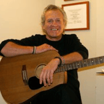 Portrait of Richie Jenkin holding an acoustic guitar and smiling.